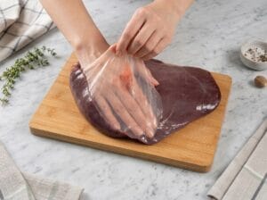 A close-up shot showing hands peeling the thin membrane off a raw beef liver