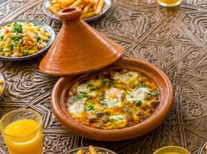 "A traditional Moroccan tagine featuring meatballs, eggs, and olives in a rich tomato sauce, served with French fries and a glass of orange juice