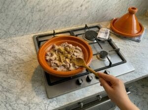 "A close-up shot showing a spoonful of ground cumin being sprinkled over beef liver cubes in a tagine