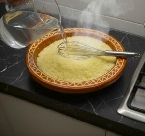 "A steaming Gasaa of couscous, accompanied by a traditional hand whisk and a bowl of cold water