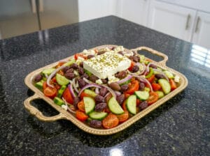 Halved tomato slices, cucumber slices, red onion slices, and green pepper slices arranged on a golden serving tray, topped with Kalamata olives and feta cheese, and drizzled with an olive oil dressing