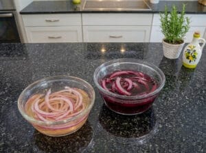 A bowl of red onion slices soaked in red wine vinegar and a bowl of red onion slices soaked in apple cider vinegar