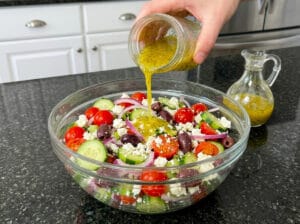 A glass bowl containing a mix of cherry tomato halves, feta cheese, Kalamata olives, cucumber slices, and thinly sliced red onions