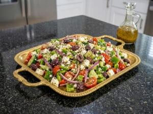 A golden serving tray containing a luxurious mix of halved cherry tomatoes, feta cheese, Kalamata olives, cucumber slices, and thin red onion slices, elegantly presented
