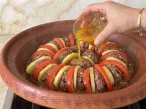 Earthenware tagine containing potato slices, kofta patties, and tomato slices neatly arranged, and a hand pouring a small cup of spices and dressing the vegetables