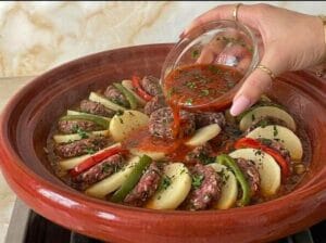 A hand pouring a little diluted sauce with spices over the kofta patties, potato slices, and green and red pepper fingers