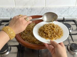 A hand scooping softened onions with a spoon from a clay tagine onto a white plate on a gas stove.